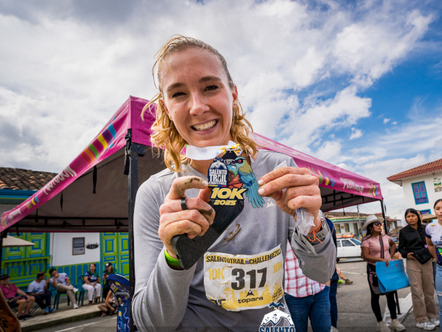 Corredora celebrando su participación en el Salento Trail Challenge con la medalla del evento, rodeada de la arquitectura tradicional de Salento.